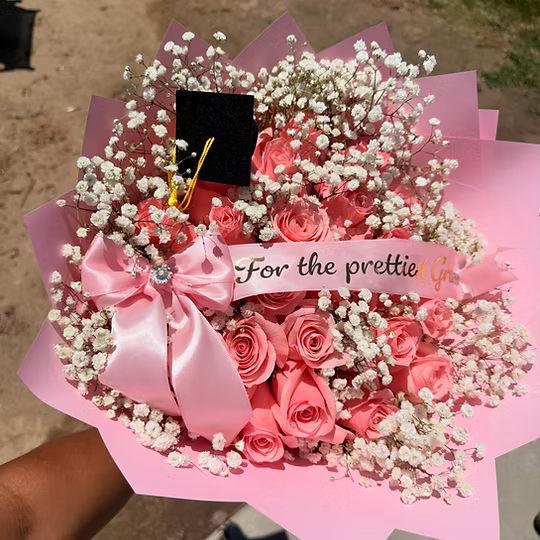 A pink rose bouquet wrapped in pink paper, filled with pink and coral roses, white baby's breath, a large pink satin bow, a small black graduation cap, and a white banner reading 'For the prettiest guy.' A hand holds the bouquet outdoors.