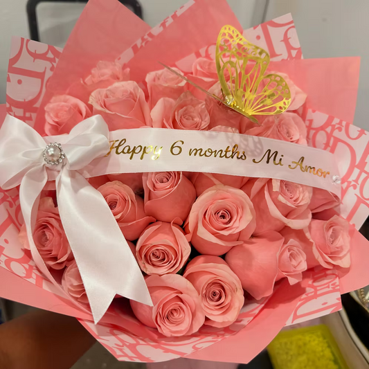 A bouquet of pink roses wrapped in pink patterned paper, tied with a white satin bow and a white ribbon that reads 'Happy 6 months Mi Amor' in gold lettering. A small gold butterfly decoration is placed on top.