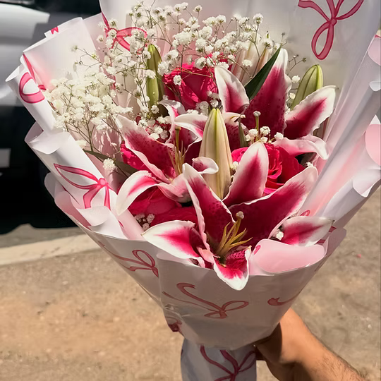 A hand holding a bouquet of lilies and baby's breath flowers wrapped in white paper with pink ribbon-like prints, photographed outdoors on a concrete surface.