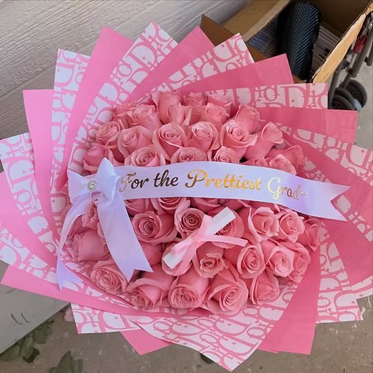 A bouquet of pink roses wrapped in pink patterned paper, with a white ribbon that reads 'For the Prettiest Grad' in gold lettering, tied with a white bow.