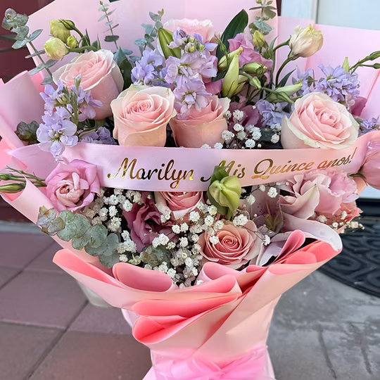 A pink and lavender floral bouquet wrapped in pink paper, featuring blush roses, purple stock flowers, eucalyptus, and a pink ribbon with gold script reading 'Marilyn 15th Quinceaño!'.
