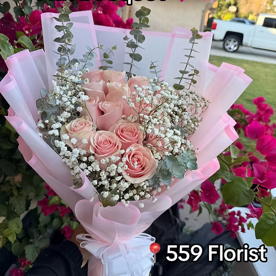 A bouquet of pink roses wrapped in light pink paper with white baby's breath and eucalyptus stems, held outdoors with flowers and a pickup truck in the background. A red heart icon and the text '559 Florist' appear near the bottom right.