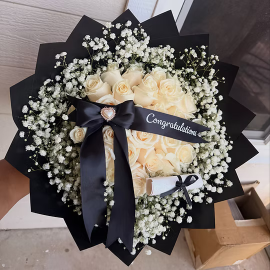 A heart-shaped bouquet of cream-colored roses wrapped in black paper with a large black ribbon bow and a 'Congratulations' banner, held by a hand.